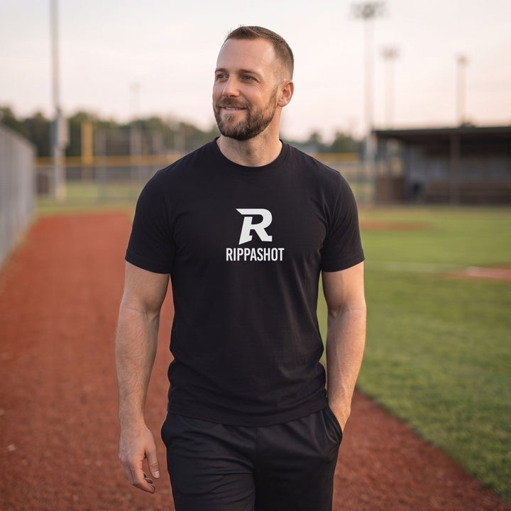 A man standing on a baseball field track wearing a black t-shirt with a white &