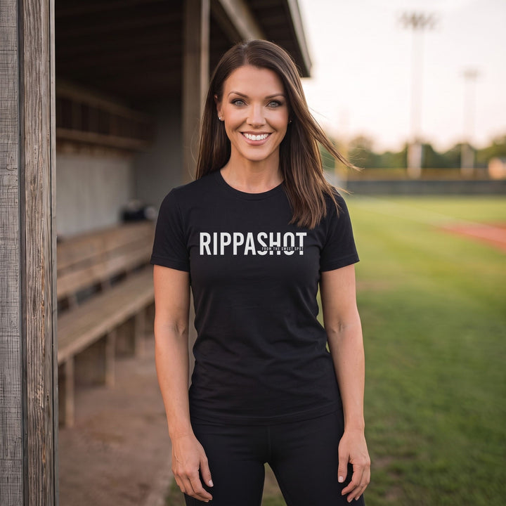 A woman stands on a baseball field wearing a black short-sleeve T-shirt with the word &