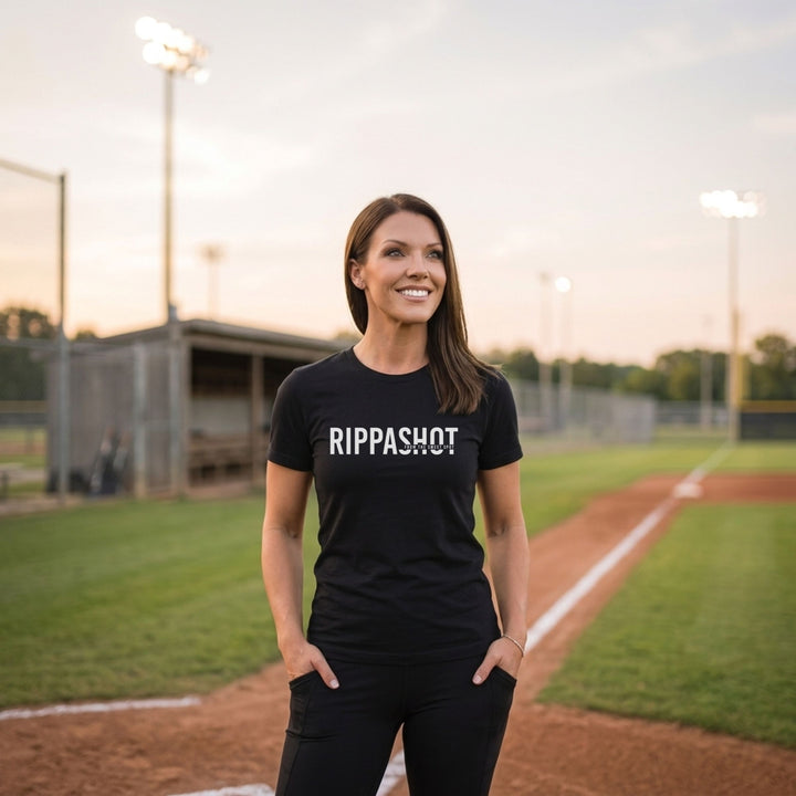 A woman stands on a baseball field wearing a black t-shirt with the text &