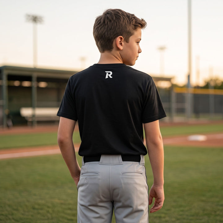A boy standing on a baseball field, viewed from behind, wearing a RIPPASHOT black short-sleeve t-shirt with a white stylized &