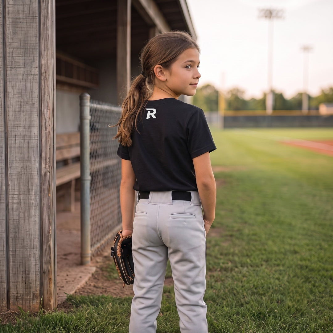 A young girl stands on a baseball field wearing a RIPPASHOT black short-sleeve t-shirt with a white letter &