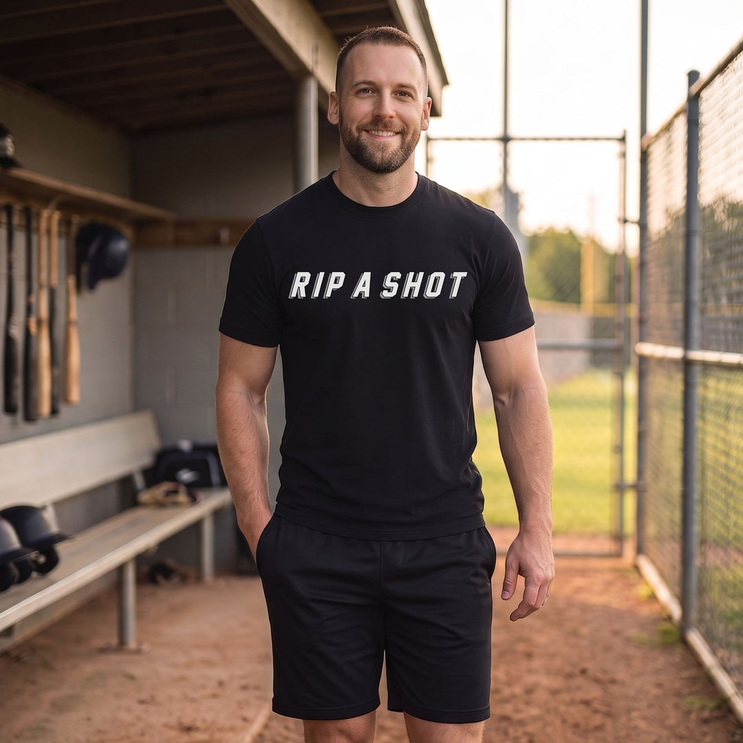 A man standing in a baseball dugout wearing a black t-shirt with 'RIP A SHOT' printed in white across the chest, paired with black athletic shorts.