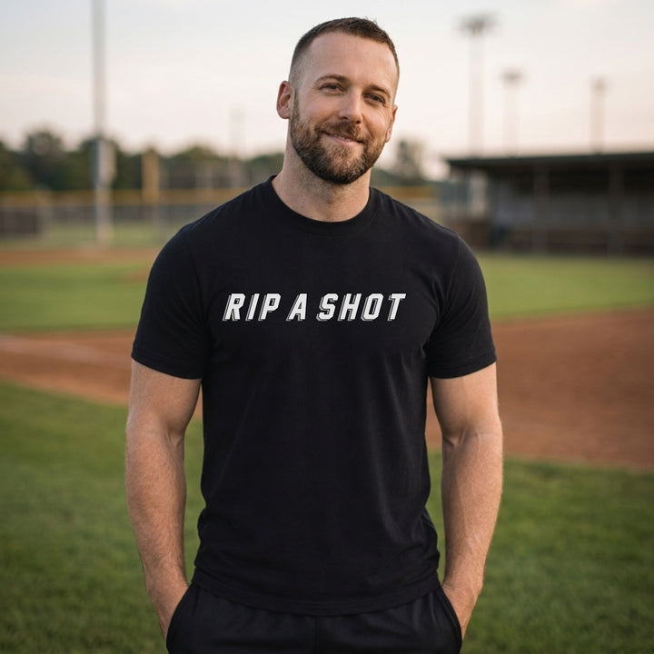A man standing on a baseball field wearing a RIPPASHOT black t-shirt with the text &