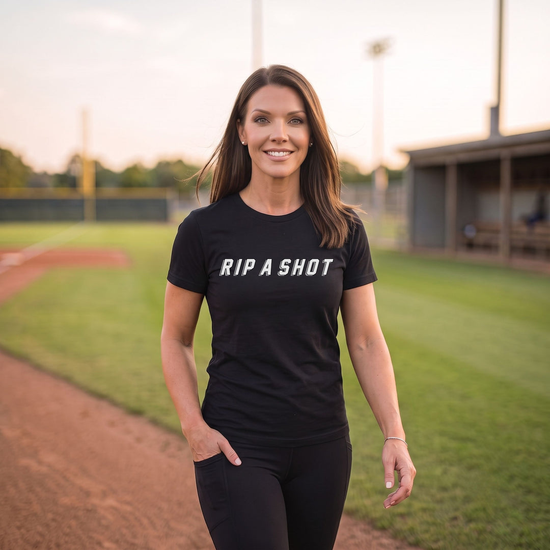 A woman stands on a baseball field at sunset wearing a black short-sleeve t-shirt with the white text 'RIP A SHOT' across the chest, paired with black leggings.