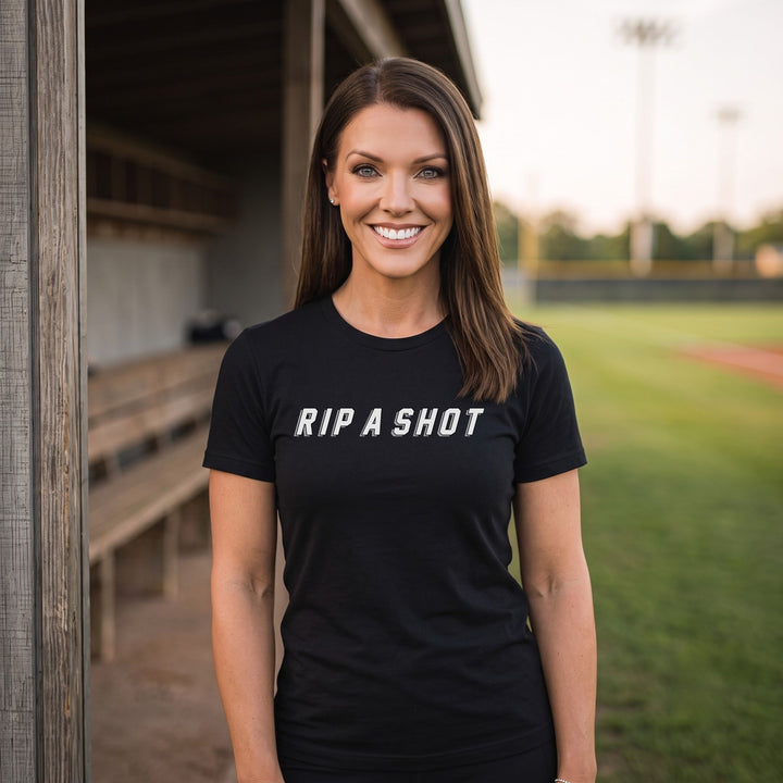 A smiling woman stands outdoors at what appears to be a baseball field, wearing a RIPPASHOT black short-sleeve T-shirt with the phrase &
