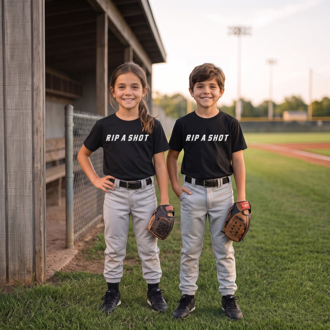 Two children standing on a baseball field wearing matching RIPPASHOT black t-shirts with 'RIP A SHOT' printed in white across the chest, paired with gray pants and holding baseball gloves.