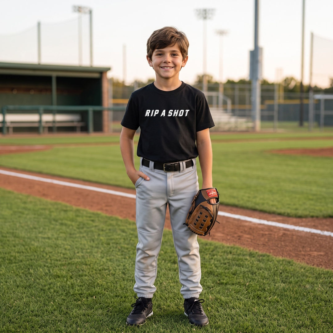 A young boy stands on a baseball field wearing a RIPPASHOT black t-shirt with the white text &