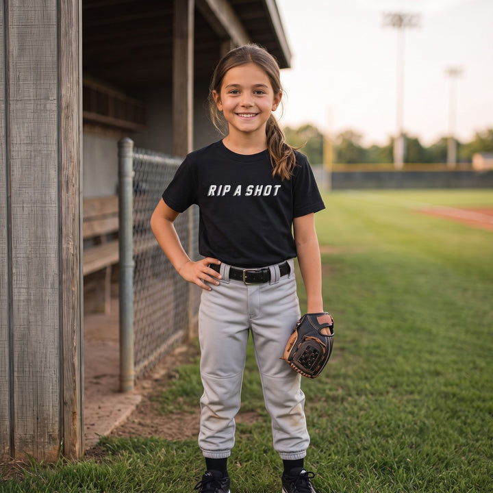 A young girl stands on a baseball field wearing a RIPPASHOT black t-shirt with the white text &