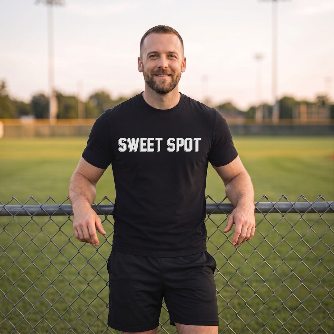 A man wearing a RIPPASHOT black t-shirt with 'SWEET SPOT' printed in white across the chest, standing behind a chain-link fence on a grassy field at sunset.