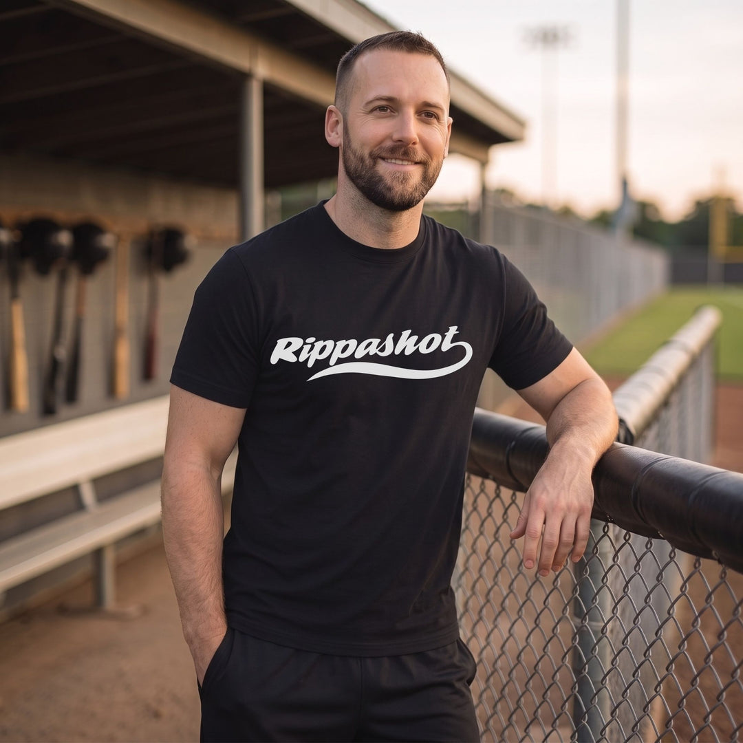 A man wearing a RIPPASHOT black short-sleeve t-shirt with a white 'Rippashot' logo, standing on a baseball field at sunset.
