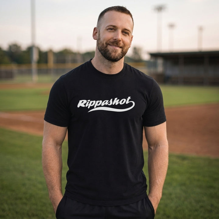 A man standing on a baseball field wearing a RIPPASHOT black t-shirt with &
