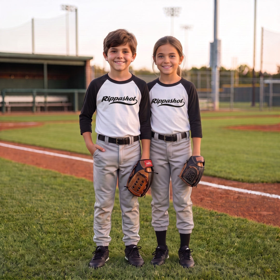 Two children standing on a baseball field wearing matching white and black raglan t-shirts with 'Rippashot' printed across the chest, paired with gray baseball pants and holding baseball gloves.