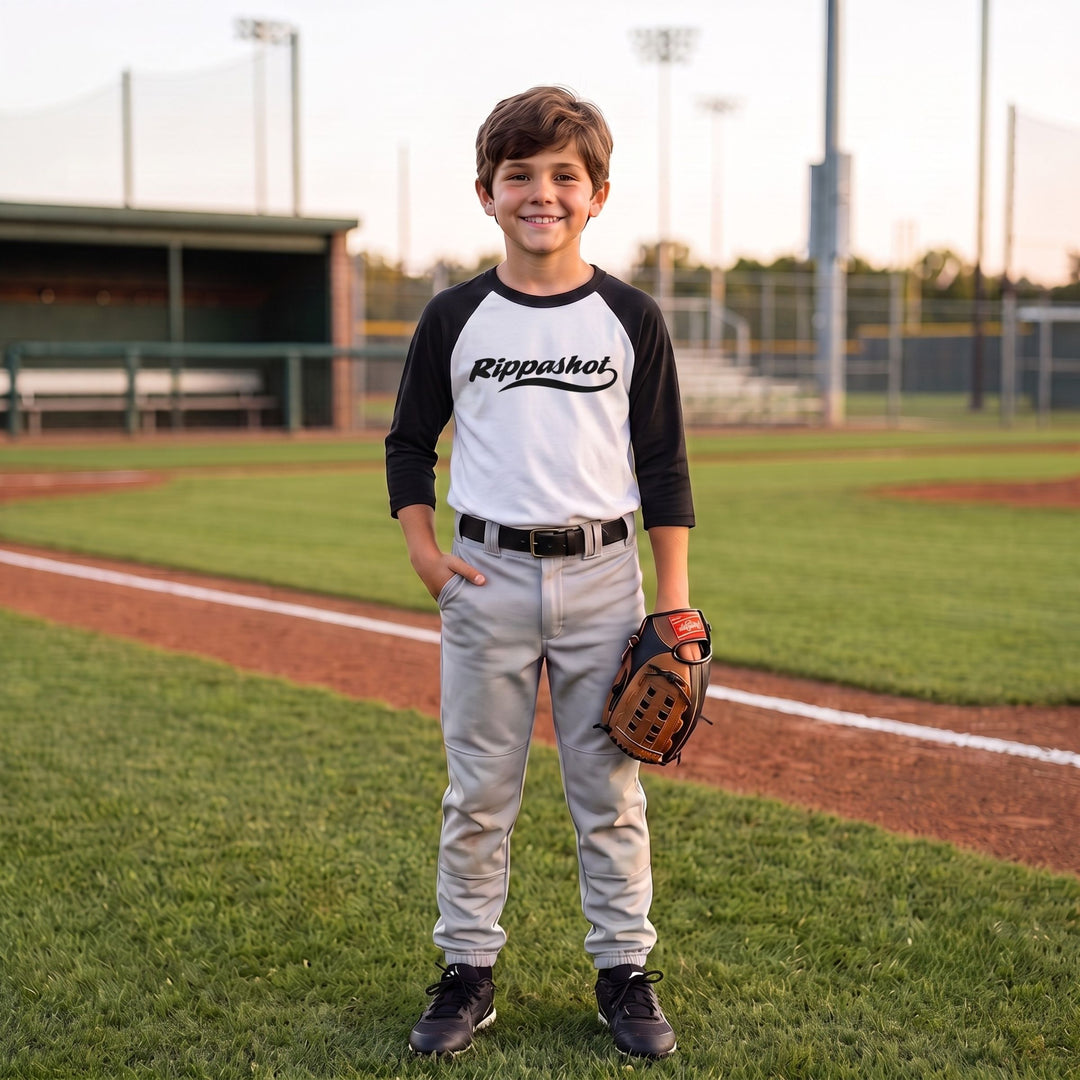 A young boy stands on a baseball field wearing a white and black raglan t-shirt with the brand name &