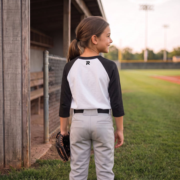 A child wearing a black and white raglan baseball t-shirt and gray baseball pants, standing on a baseball field near a dugout.