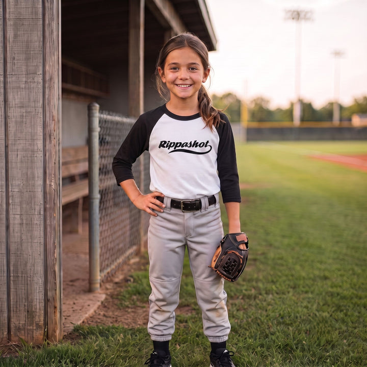 A young girl stands on a baseball field wearing a white and black raglan baseball jersey with &