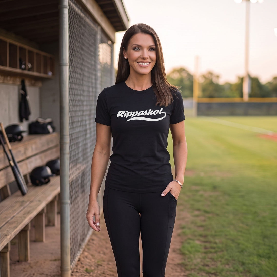 A woman stands on a baseball field wearing a black short-sleeve t-shirt with 'Rippashot' printed in white script across the chest, paired with black leggings.