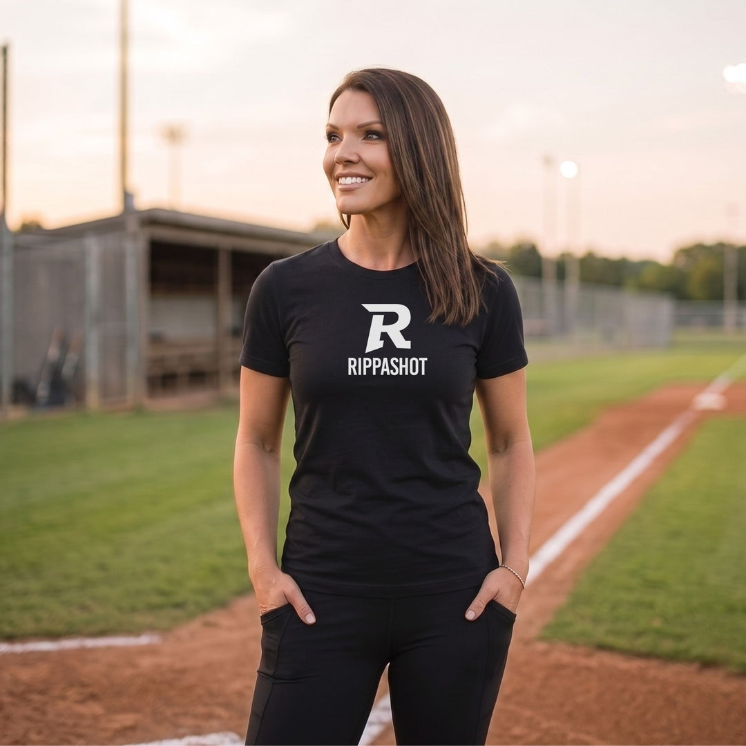 A woman stands on a baseball field wearing a black short-sleeve t-shirt with a white 'R' logo and the word 'RIPPASHOT' printed on the chest, paired with black pants.