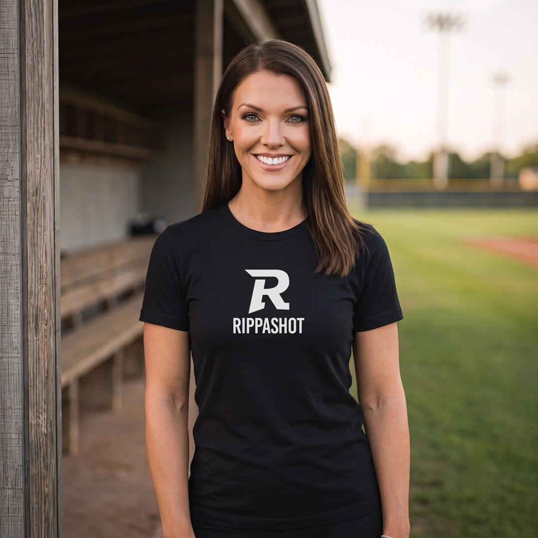 A smiling woman stands outdoors on a baseball field wearing a RIPPASHOT black short-sleeve T-shirt with a large white stylized letter R and the word RIPPASHOT printed on the front.