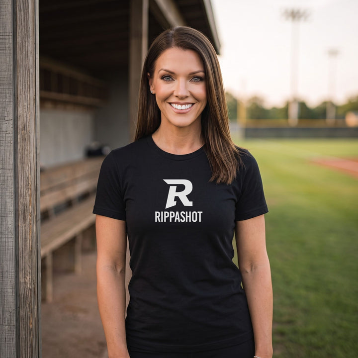 A smiling woman stands outdoors on a baseball field wearing a RIPPASHOT black short-sleeve T-shirt with a large white stylized letter R and the word RIPPASHOT printed on the front.