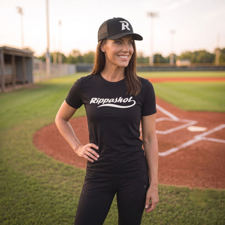 A woman stands on a baseball field wearing a black short-sleeve t-shirt with &