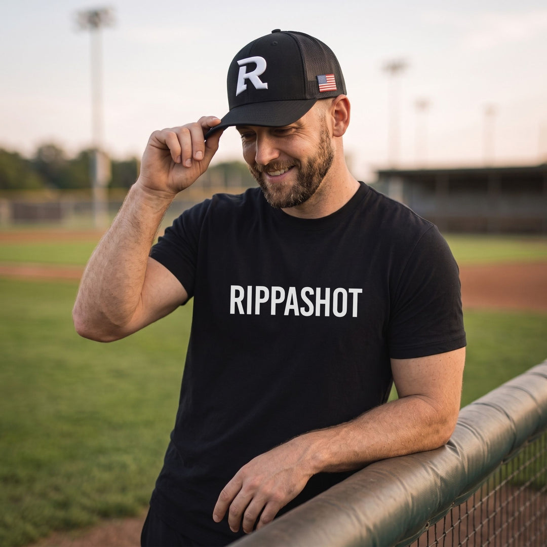 A man wearing a black baseball cap with a white 'R' logo and a black t-shirt with 'RIPPASHOT' printed in white, standing on a baseball field at sunset.
