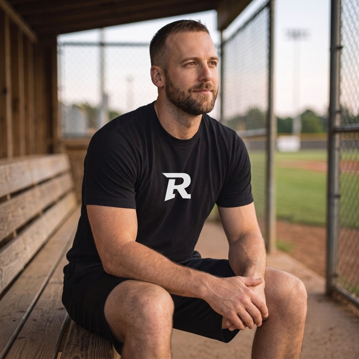 A man sitting on a wooden bench in a baseball dugout wearing a RIPPASHOT black t-shirt with a white stylized &