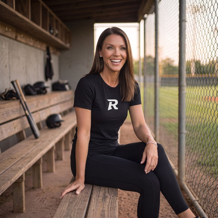A woman sitting on a wooden bench in a baseball dugout wearing a RIPPASHOT black t-shirt with a white letter &